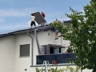 Bild von zwei Männer auf einem Dach, die ein Solarpanel tragen. An der Seite sieht man, dass sie eine Leiter auf einen Anbau gestellt haben, um aufs Dach zu kommen. – Bildquelle: Michael Silbermann 