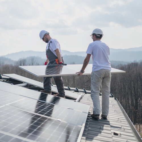 Ungesicherter Dachinstallation einer Photovoltaikanlage Negativbeispiel - Zwei ungesicherte Bauarbeiter transportieren ein Solarmodul auf einem Dach an einer Absturzkante.