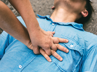 Bild eines Mannes mit blauem Hemd, bei dem eine Herzdruck-Massage durchgeführt wird. – Bildquelle: PlatooStock - stock.adobe.com